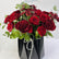 A bouquet of deep red roses with green foliage arranged in a black geometric paper vase tied with a white ribbon, photographed against a plain light background.