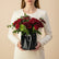 A bouquet of deep red roses and green eucalyptus stems arranged in a black faceted geometric vase, tied with a white string, displayed on a white table against a light gray background.