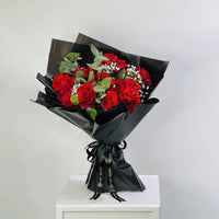 A bouquet of red roses wrapped in black paper with green leaves and white baby's breath, tied with a black ribbon, displayed on a white pedestal against a light gray background.