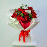 A bouquet of red roses wrapped in white paper with a red ribbon bow, displayed on a white pedestal against a plain light background.