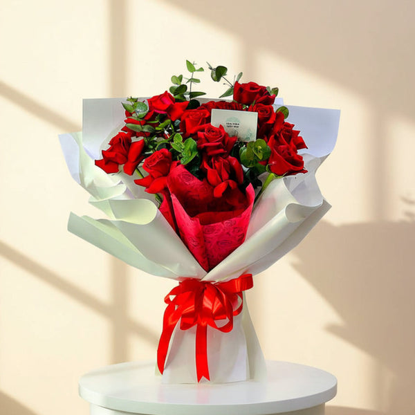 A bouquet of red roses wrapped in white paper with a red ribbon bow, displayed on a white pedestal against a plain light background.