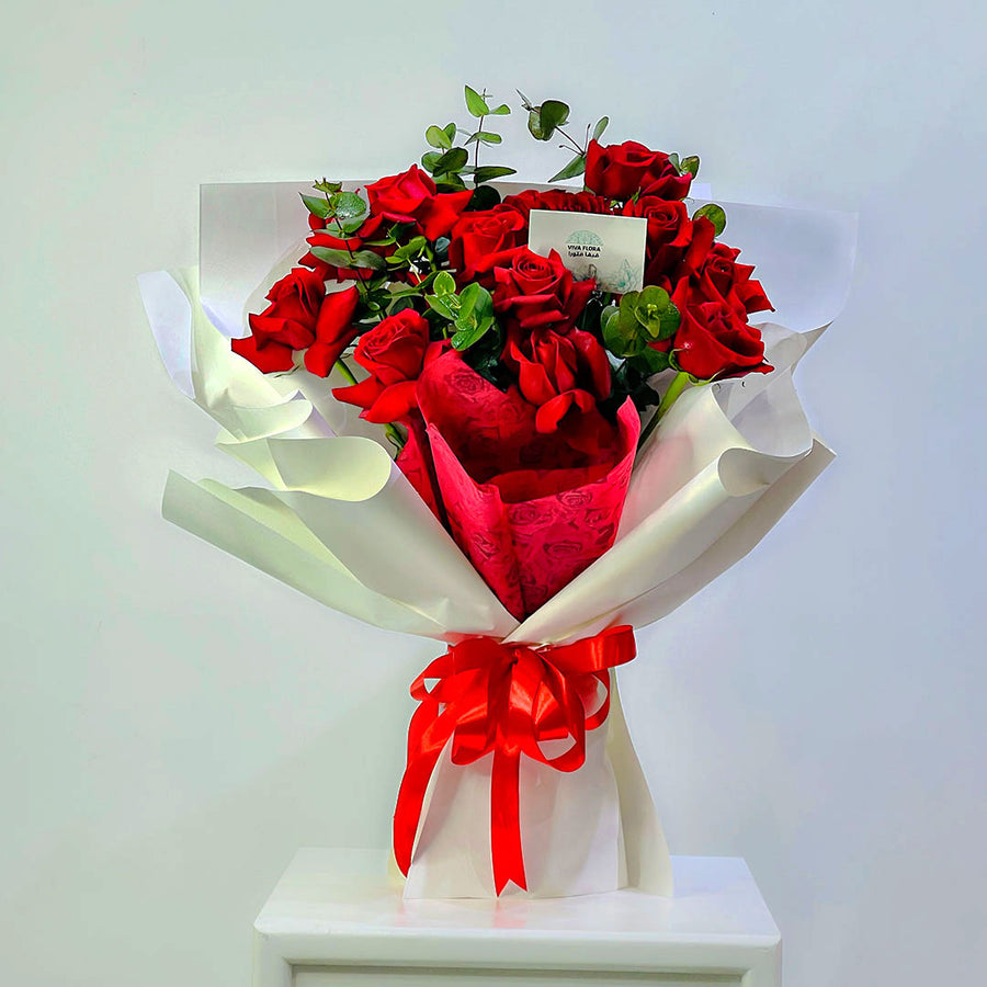A bouquet of red roses wrapped in white paper with a red ribbon bow, displayed on a white pedestal against a plain light background.