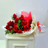 A bouquet of red roses wrapped in white and red tissue paper, displayed on a white pedestal against a plain light background.