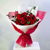 A bouquet of red roses wrapped in white and bright pink patterned paper, tied with a matching bow, displayed on a white pedestal against a light gray background. flower delivery in qatar