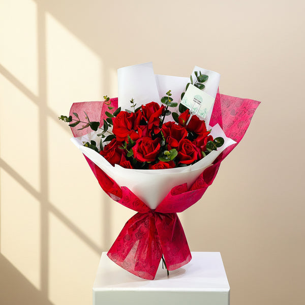 A bouquet of red roses wrapped in white and bright pink patterned paper, tied with a matching bow, displayed on a white pedestal against a light gray background. flower delivery in qatar
