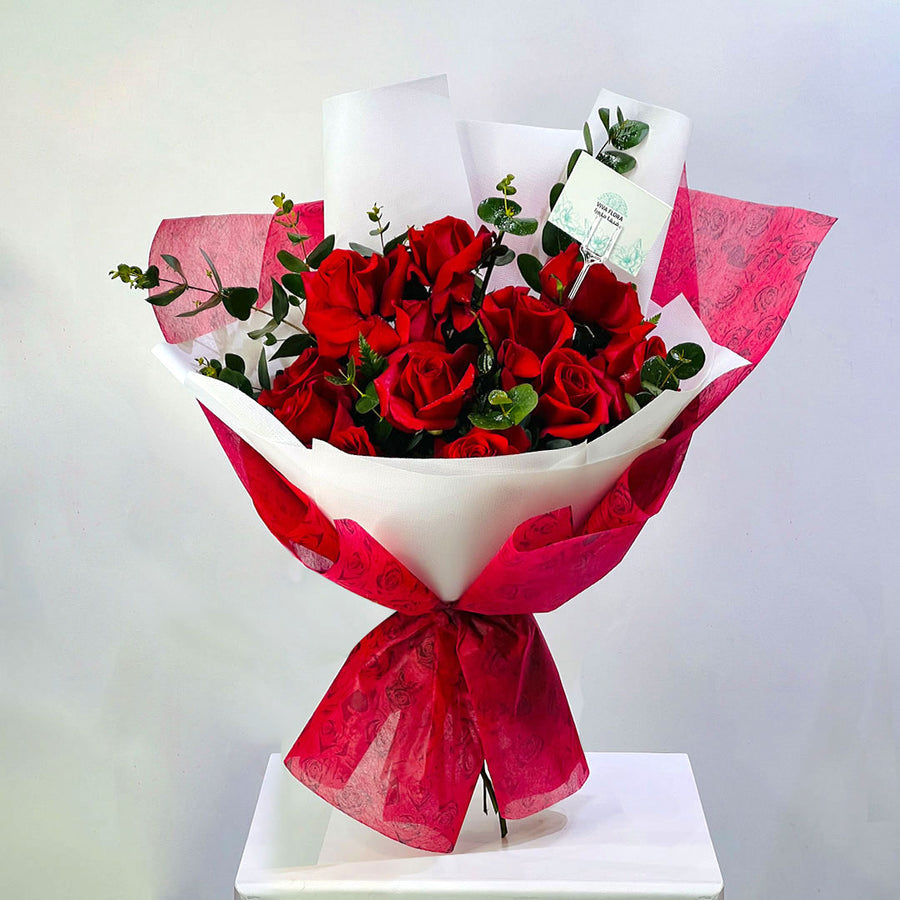 A bouquet of red roses wrapped in white and bright pink patterned paper, tied with a matching bow, displayed on a white pedestal against a light gray background. flower delivery in qatar
