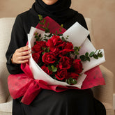 A bouquet of red roses wrapped in white and bright pink patterned paper, tied with a matching bow, displayed on a white pedestal against a light gray background.