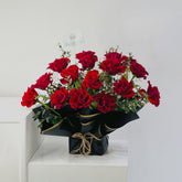 A bouquet of deep red roses wrapped in black paper with gold ribbon accents, displayed on a white pedestal against a plain light gray background.