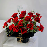 A vibrant bouquet of approximately 20 red roses with green leaves and small white filler flowers, wrapped in black paper with a gold ribbon and a small card, displayed on a white pedestal.