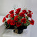 A vibrant bouquet of approximately 20 red roses with green leaves and small white filler flowers, wrapped in black paper with a gold ribbon and a small card, displayed on a white pedestal.