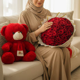 A red plush teddy bear with a white snout and a red ribbon sits beside a large bouquet of red roses wrapped in white paper with gold trim, displayed on white pedestals against a beige wall with soft window shadows.