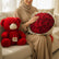 A red plush teddy bear with a white snout and a red ribbon sits beside a large bouquet of red roses wrapped in white paper with gold trim, displayed on white pedestals against a beige wall with soft window shadows.