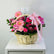 A floral arrangement in a woven wicker basket with a pink ribbon bow, displayed on a white cabinet against a light gray wall.