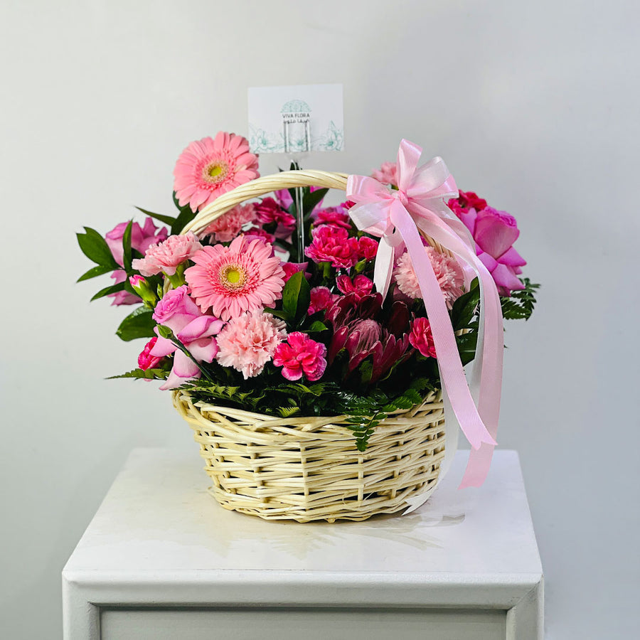 A floral arrangement in a woven wicker basket with a pink ribbon bow, displayed on a white cabinet against a light gray wall.