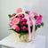 A wicker basket filled with a colorful floral arrangement featuring pink gerbera daisies, pink roses, pink carnations, burgundy protea, and green foliage, tied with a large pink ribbon bow. A small card is visible behind the bouquet, and the arrangement sits on a white surface against a light gray background.
