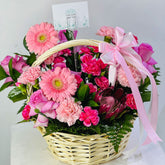 A wicker flower basket filled with pink and red flowers, green foliage, and a light pink ribbon bow, with a small card visible at the top.