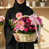 A floral arrangement in a woven wicker basket with a pink ribbon bow, displayed on a white cabinet against a light gray wall.