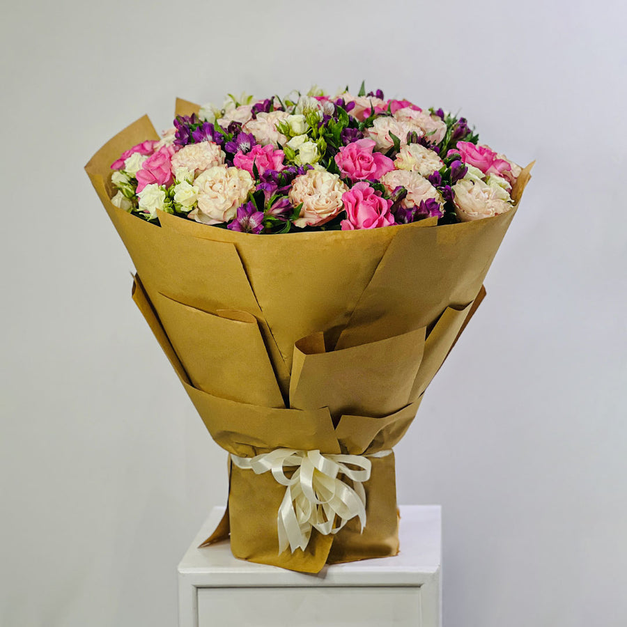 A large bouquet of pink, cream, and purple flowers wrapped in brown kraft paper and tied with a cream ribbon, displayed on a white pedestal against a light gray background.