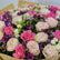 A close-up of a wrapped flower bouquet filled with pink and cream roses, cream peonies, purple alstroemeria, and green foliage, wrapped in brown kraft paper.
