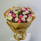 A large bouquet of pink, purple, and cream carnations wrapped in brown paper and tied with a cream ribbon, displayed on a white pedestal against a light gray background.