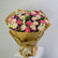 A large bouquet of pink, purple, and cream carnations wrapped in brown paper and tied with a cream ribbon, displayed on a white pedestal against a light gray background.