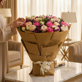 A large bouquet of pink, cream, and purple flowers wrapped in brown kraft paper and tied with a cream ribbon, displayed on a white pedestal against a light gray background.