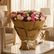 A large bouquet of pink, cream, and purple flowers wrapped in brown kraft paper and tied with a cream ribbon, displayed on a white pedestal against a light gray background.