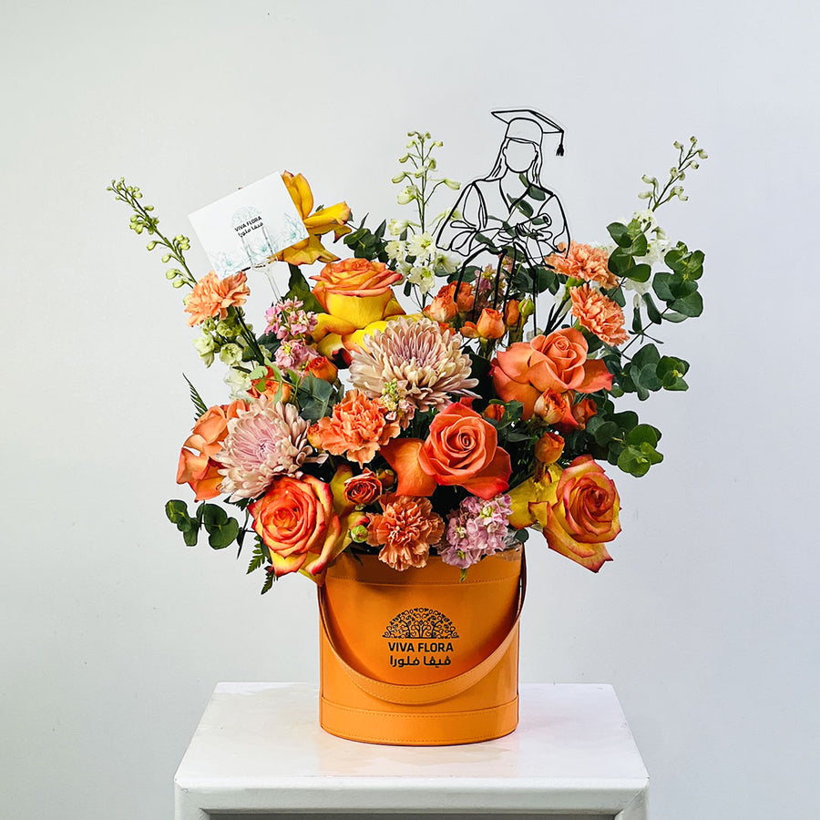 A vibrant graduation floral arrangement in an orange cylindrical bucket box, displayed on a white pedestal against a light gray background. The bouquet features orange and yellow roses, pink chrysanthemums, green foliage, and a black metal graduate silhouette decoration.