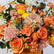 A vibrant floral arrangement featuring orange and yellow roses, pink and peach chrysanthemums, and orange carnations with green foliage, set against a light gray background.