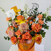 A graduation-themed floral arrangement in a metallic orange bucket with the brand name 'VIVA FLORA' printed on it. The bouquet features orange and yellow roses, pink and orange carnations, purple chrysanthemums, green eucalyptus, and a black wire silhouette of a graduate wearing a cap.