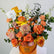 A graduation-themed floral arrangement in a metallic orange bucket with the brand name 'VIVA FLORA' printed on it. The bouquet features orange and yellow roses, pink and orange carnations, purple chrysanthemums, green eucalyptus, and a black wire silhouette of a graduate wearing a cap.
