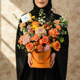 A vibrant graduation floral arrangement in an orange cylindrical bucket box, displayed on a white pedestal against a light gray background. The bouquet features orange and yellow roses, pink chrysanthemums, green foliage, and a black metal graduate silhouette decoration.