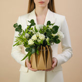 A floral arrangement of white roses, lilies, and green eucalyptus stems in a brown geometric paper bag vase, displayed on a white table against a light gray background.