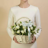 A white rose and eustoma flower basket in a woven wicker basket, tied with a white ribbon printed with the name 'VIVA EBRA', displayed on a white surface against a light gray background.