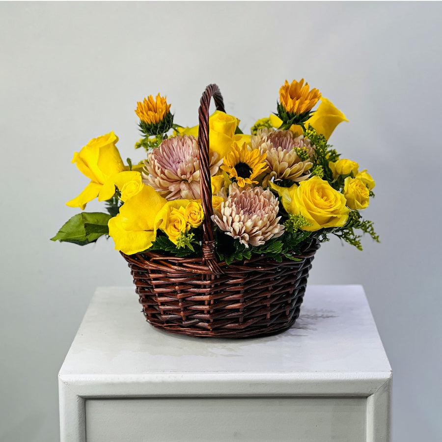 A wicker flower basket filled with yellow roses, sunflowers, and pale pink chrysanthemums, displayed on a white pedestal against a light gray background.