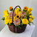A wicker flower basket filled with yellow roses, chrysanthemums, sunflowers, and green foliage, displayed on a white surface against a light gray background.