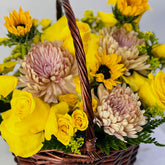 A wicker flower basket filled with bright yellow roses, yellow sunflowers, pale pink chrysanthemums, and green foliage, photographed close up against a light background.