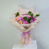 A colorful flower bouquet wrapped in light beige tissue paper and tied with a lavender ribbon, featuring pink, purple, and peach roses with green leaves and a 'Thank You' card, displayed on a white pedestal against a light gray background.