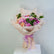 A colorful flower bouquet wrapped in light beige tissue paper and tied with a lavender ribbon, featuring pink, purple, and peach roses with green leaves and a 'Thank You' card, displayed on a white pedestal against a light gray background.