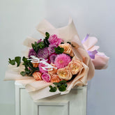 A floral bouquet with pink and peach roses, purple chrysanthemums, green eucalyptus leaves, and a white 'Happy Birthday' card, wrapped in beige tissue paper and tied with a lavender ribbon, displayed on a white cabinet against a light gray wall.