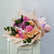 A floral bouquet with pink and peach roses, purple chrysanthemums, green eucalyptus leaves, and a white 'Happy Birthday' card, wrapped in beige tissue paper and tied with a lavender ribbon, displayed on a white cabinet against a light gray wall.