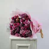 A large bouquet of pink and purple roses wrapped in sheer pink tissue paper, displayed on a white cabinet against a plain light gray wall.
