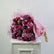 A large bouquet of pink and purple roses wrapped in sheer pink tissue paper, displayed on a white cabinet against a plain light gray wall.