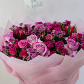 A vibrant bouquet of pink and purple roses with green foliage, wrapped in light pink tissue paper. A small card with the brand name 'VIVA FLORA' is visible at the top.