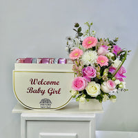 A floral welcome gift arrangement featuring a cream-colored box with the text 'Welcome Baby Girl' and pink wrapped items peeking out from behind, next to a vibrant bouquet of pink, white, and purple flowers in a basket, displayed on a white cabinet against a light gray wall.