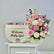 A floral welcome gift arrangement featuring a cream-colored box with the text 'Welcome Baby Girl' and pink wrapped items peeking out from behind, next to a vibrant bouquet of pink, white, and purple flowers in a basket, displayed on a white cabinet against a light gray wall.