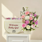 A floral welcome gift arrangement featuring a cream-colored box with the text 'Welcome Baby Girl' and pink wrapped items peeking out from behind, next to a vibrant bouquet of pink, white, and purple flowers in a basket, displayed on a white cabinet against a light gray wall.