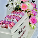 A white baby shower favor box filled with pink and purple mosaic tile favors, decorated with a 'Welcome Baby Girl' sign, surrounded by a colorful bouquet of pink and white flowers.