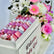 A white baby shower favor box filled with pink and purple mosaic tile favors, decorated with a 'Welcome Baby Girl' sign, surrounded by a colorful bouquet of pink and white flowers.