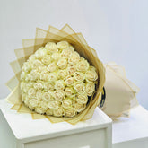 A large bouquet of cream-colored roses wrapped in sheer beige tissue paper, displayed on a white pedestal against a light gray background.